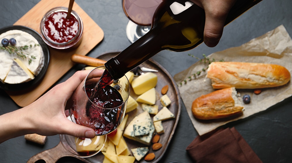 Pouring red wine in a glass over cheese platter and bread at the top wine bar